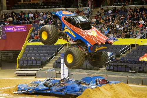 A monster truck named "Superman" jumps over crushed cars at a lively arena event, with a cheering crowd in the background.