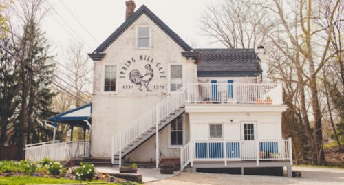 A charming white building with a sign reading "Spring Mill Café," featuring a chicken illustration and a staircase.
