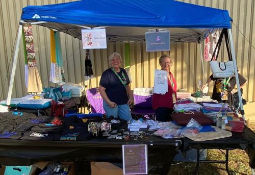 Two women smile behind a vendor booth filled with various crafts and merchandise under a blue canopy.