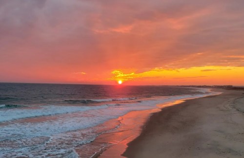 A serene beach at sunset, with vibrant orange and pink hues reflecting on the water and gentle waves lapping the shore.