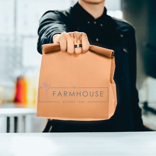 A person holding a brown takeout bag with "The Farmhouse Kitchen + Bar" logo in a kitchen setting.