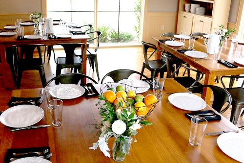 A dining room with wooden tables set for a meal, featuring plates, glasses, and a centerpiece of fruit and flowers.