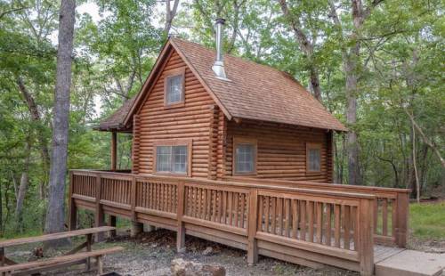 A cozy log cabin surrounded by trees, featuring a wooden deck and a sloped roof.