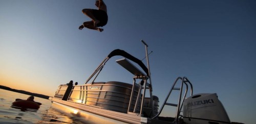 A person jumps off a boat into the water at sunset, with a calm lake and a motorboat in the background.