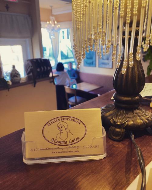 A business card for Mamma Luisa Italian Restaurant on a table near a lamp, with a dining area in the background.