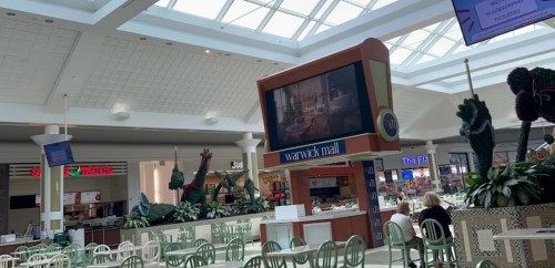 A spacious mall interior with a large screen, seating areas, and decorative plants under a glass ceiling.