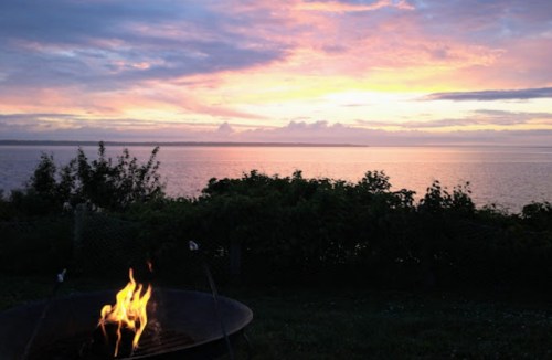 A serene sunset over the water, with vibrant colors in the sky and a small fire pit in the foreground.
