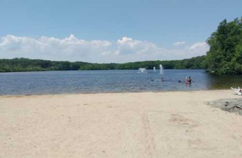 A sandy beach by a calm lake with people swimming and fountains in the background under a clear blue sky.