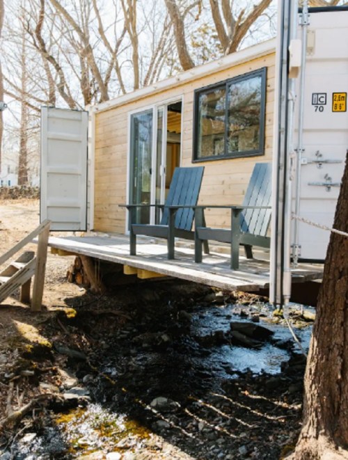 A modern wooden cabin with a deck overlooking a small stream, surrounded by trees in a serene outdoor setting.