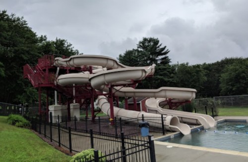 Water slides at a pool surrounded by greenery, with a cloudy sky overhead.
