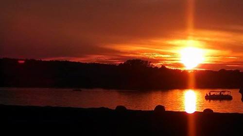 A vibrant sunset over a calm lake, with silhouettes of trees and a boat on the water.
