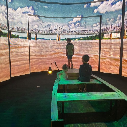 Two children sit in a boat, facing a large screen displaying a river and bridge under a blue sky.