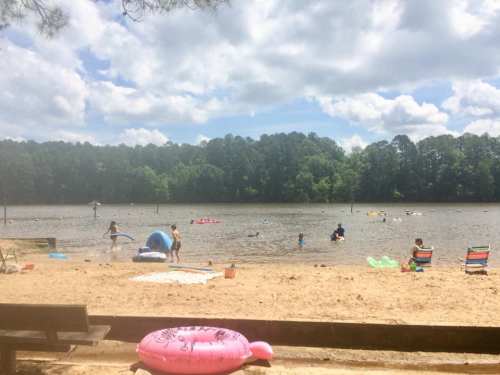 A sandy beach by a lake with people swimming, lounging, and enjoying the sun under a partly cloudy sky.