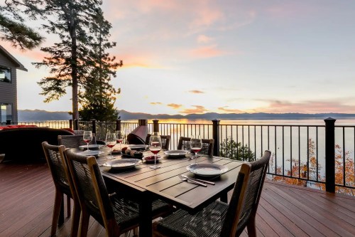 A dining table set for a meal on a deck, overlooking a serene lake at sunset with mountains in the background.