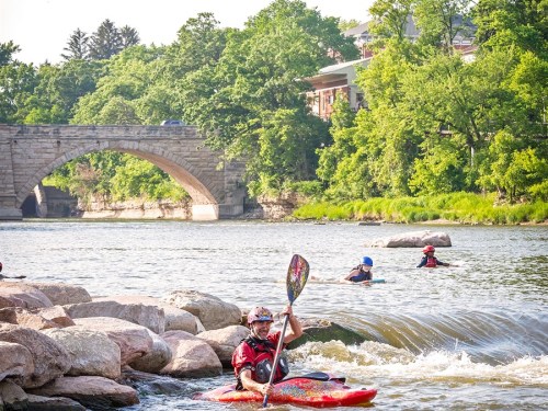 A person in a colorful kayak paddles on a river, with a stone bridge and trees in the background.
