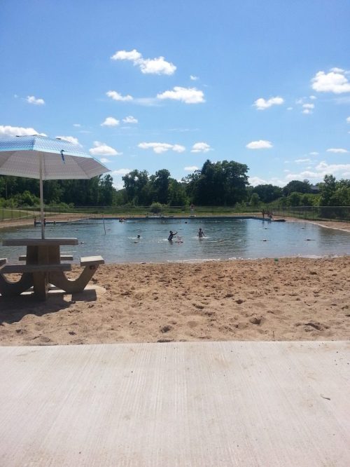 A sandy area by a swimming pond with people enjoying the water under a clear blue sky and fluffy clouds.