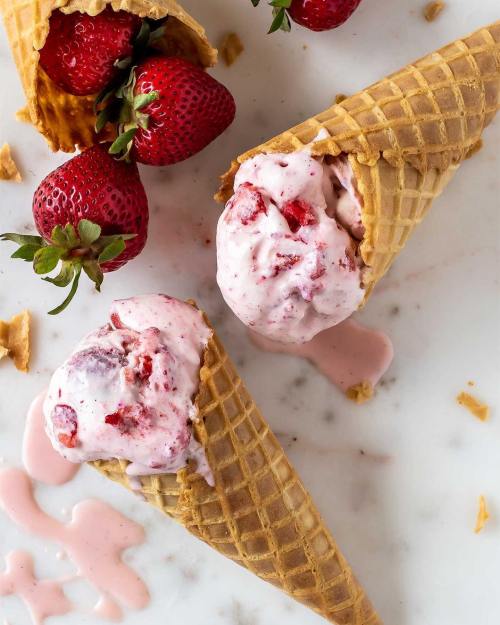 Two waffle cones filled with strawberry ice cream, surrounded by fresh strawberries and melted ice cream on a marble surface.