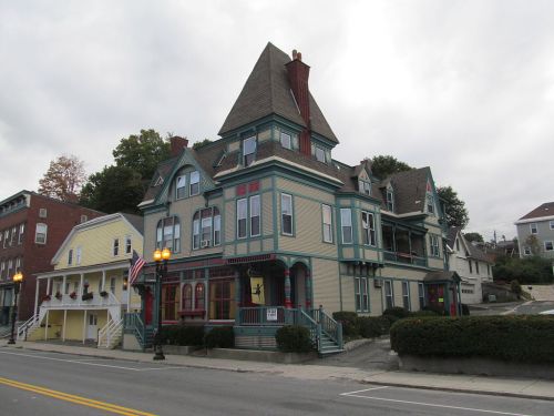 A colorful Victorian-style house with a steep roof, surrounded by greenery and other buildings on a cloudy day.