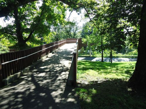 A wooden ramp leads through a shaded park, surrounded by trees and greenery. Sunlight filters through the leaves.