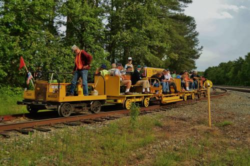A group of people riding on a yellow rail car along a tree-lined track on a sunny day.