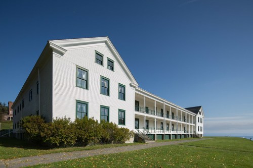 A large white building with green trim, featuring a porch and surrounded by grass, overlooking a body of water.
