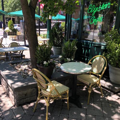A cozy outdoor café setting with a small table, two chairs, and greenery under a tree, featuring a neon sign.
