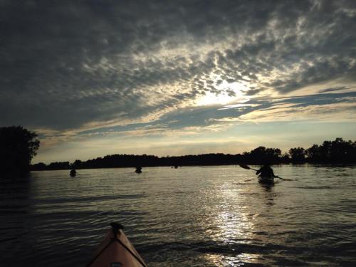 A serene sunset over a calm lake, with kayakers paddling in the foreground and dramatic clouds in the sky.