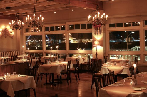 Cozy restaurant interior with candlelit tables, chandeliers, and a view of city lights through large windows.
