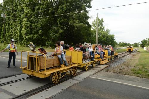 A group of people rides on a yellow train-like vehicle along a railway, with a conductor guiding them.