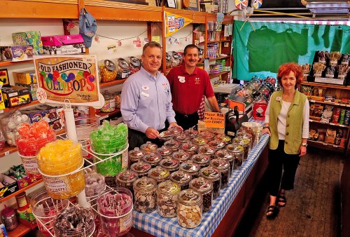 Three people stand behind a colorful candy counter filled with jars of sweets in a vintage-style shop.
