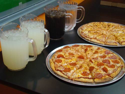 Two pizzas with pepperoni slices, accompanied by pitchers of lemonade and soda on a black table.