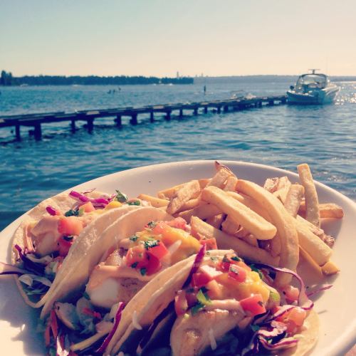 A plate of fish tacos with salsa and fries, with a scenic view of a lake and a boat in the background.
