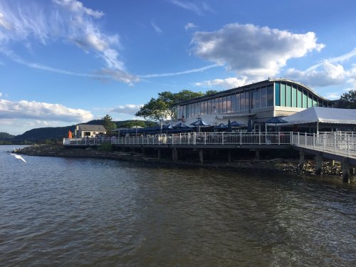 A waterfront restaurant with outdoor seating, surrounded by trees and a blue sky with clouds.
