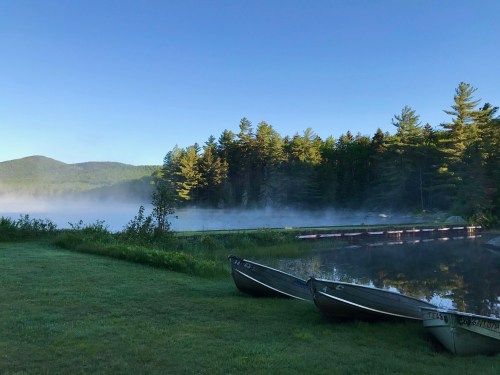 Serene lake scene at dawn with mist rising, surrounded by trees and two boats on the shore.