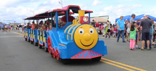 A colorful children's train ride with smiling kids, moving through a lively outdoor event with crowds in the background.