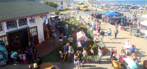 A bustling outdoor festival with people enjoying activities, food stalls, and decorations, near a beach and parking area.