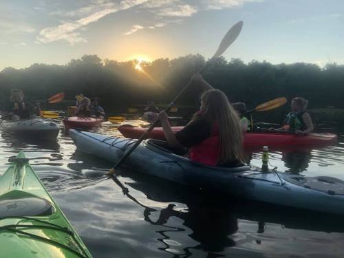 A group of people kayaking at sunset, with reflections on the water and lush trees in the background.