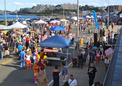 A bustling outdoor festival with crowds, tents, and a scenic coastal backdrop. People are enjoying various activities.