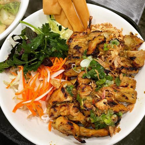 A plate of grilled chicken with fresh herbs, pickled vegetables, and crispy spring rolls.