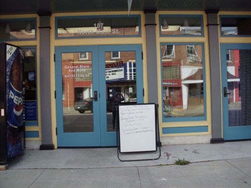 Entrance of a general store with large glass doors and a signboard displaying information outside.