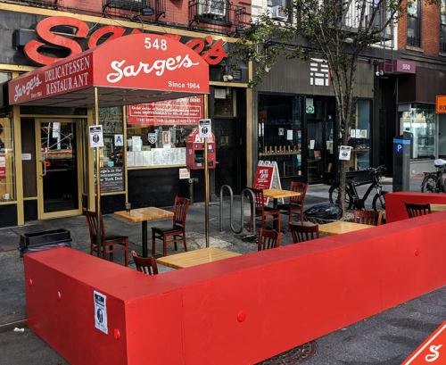 Exterior of Sarge's Delicatessen with red seating area and tables, located on a city street.