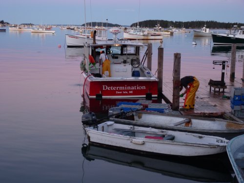 A fishing boat named "Determination" docked at a harbor with other boats and calm water at sunset.