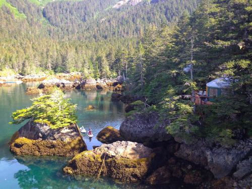 A serene coastal scene with rocky shores, lush trees, and two people kayaking in calm waters.