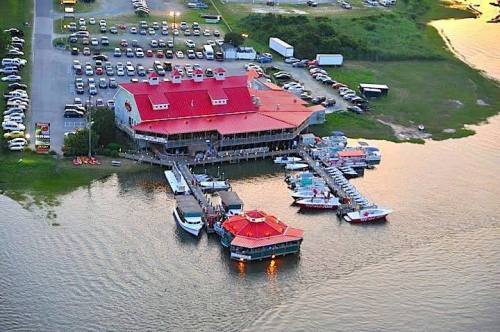 Aerial view of a waterfront restaurant with a red roof, surrounded by boats and parked cars near a calm waterway.