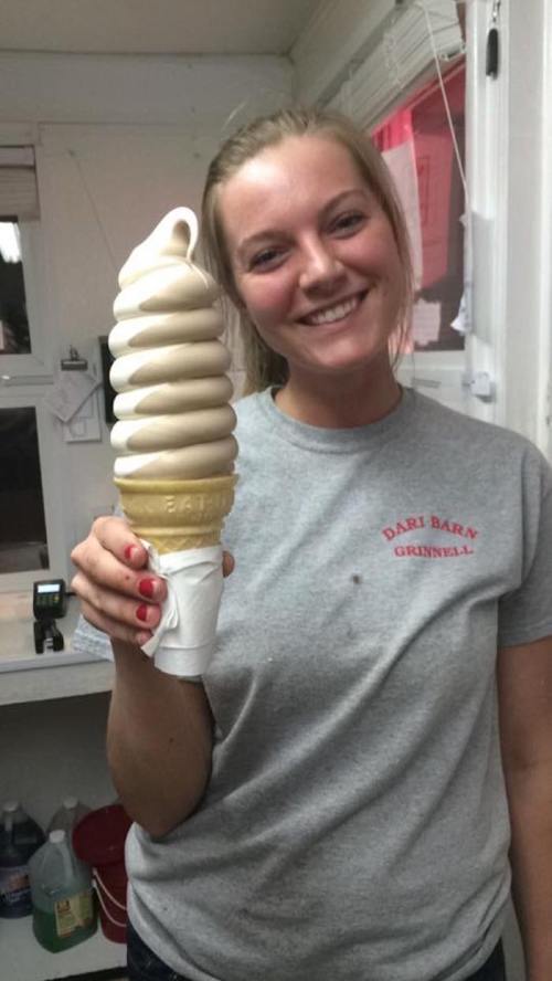 A smiling woman holds a large swirl ice cream cone in a shop, wearing a gray Dari Barn t-shirt.