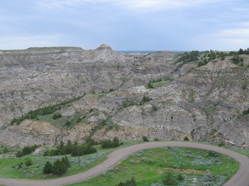 A winding road curves through rugged, hilly terrain with sparse greenery under a cloudy sky.