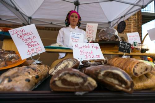 A baker stands behind a table displaying various breads with price signs, including Irish oatmeal raisin and marbled rye.