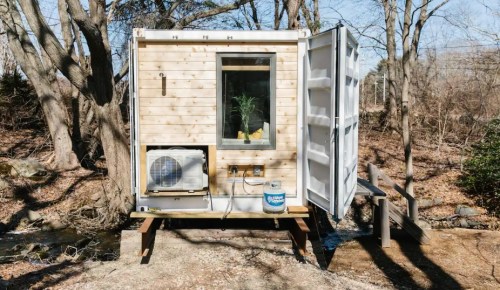 A small wooden cabin with a large window, set in a wooded area near a stream, featuring an air conditioning unit.
