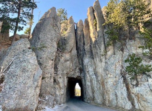 A narrow road passes through a tall rock formation with jagged peaks and pine trees in the background.