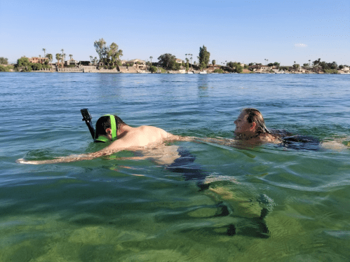 Two people swimming in clear water, one using a snorkel and the other swimming beside them, with a sunny backdrop.